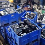 Heavy-duty blue industrial recycling bins stacked with bulk corporate UPS batteries and scrap lead-acid power cells at a warehouse yard